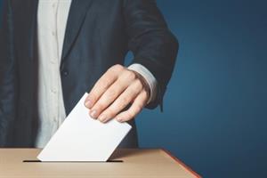 Mann im dunkelblauen Sakko wirft weißen Zettel in Wahlurne aus hellem Holz, dahinter blauer Hintergrund